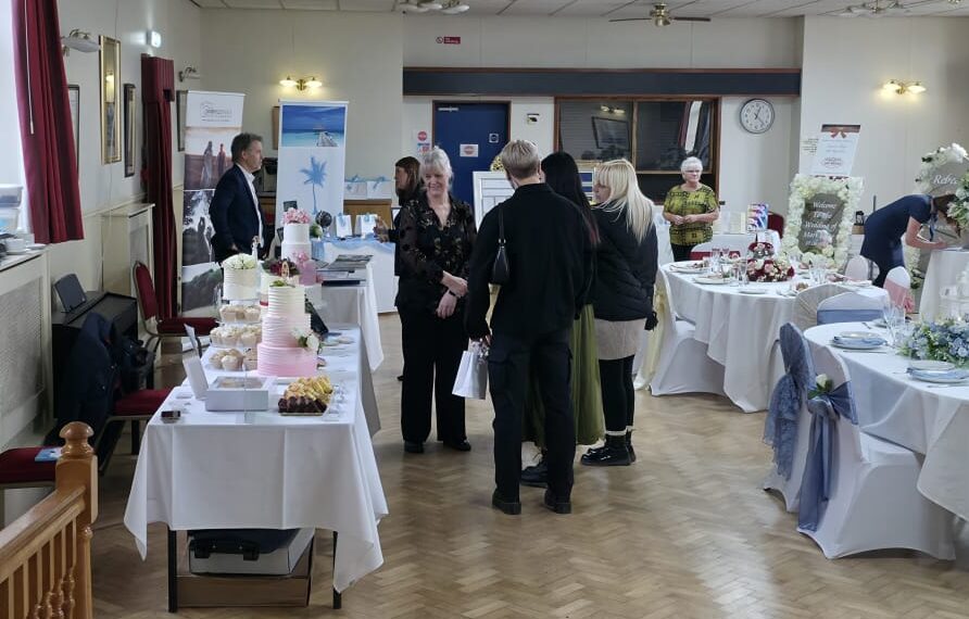 Visitors browsing wedding supplier exhibition stands at a wedding fayre, with displays and decorated tables.